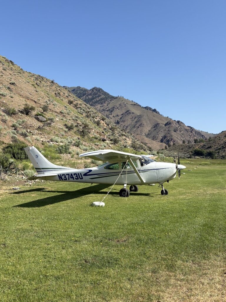 A small white single-engine airplane is parked on a grassy field near hills under a clear blue sky.