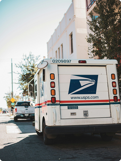 A USPS mail delivery truck parked on a city street, with buildings and trees visible in the background.