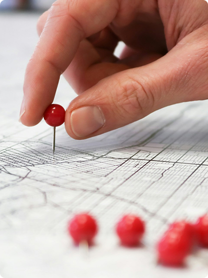 A hand places a red pushpin on a map, with several other red pins lying nearby on the map’s surface.