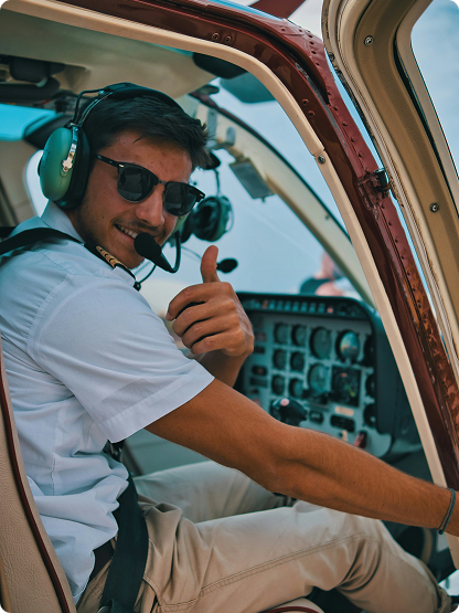 A pilot wearing a headset and sunglasses sits in the cockpit of a small aircraft, giving a thumbs up while looking toward the camera.