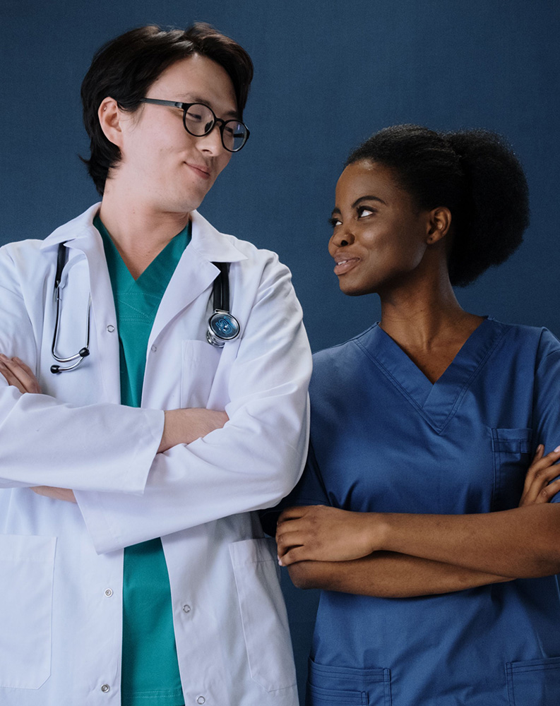 Two medical professionals in scrubs and lab coats stand side by side with arms crossed, looking at each other and smiling against a plain blue background.