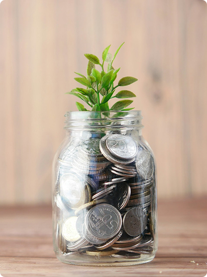 A glass jar filled with coins has a small green plant growing from the top, set against a plain wooden background.